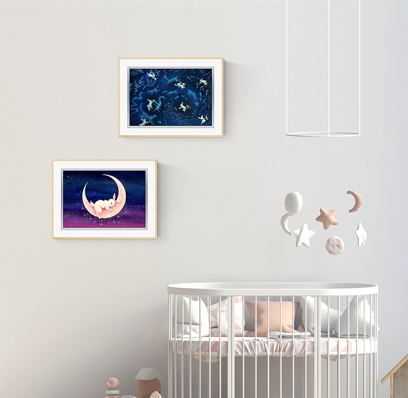 May include: Two framed illustrations of a white rabbit sleeping on a crescent moon and a constellation of stars in a dark blue sky. A white crib with a pink and white patterned bedding set is in the foreground.