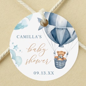 May include: A round baby shower favor tag with a blue and white hot air balloon, a teddy bear, and clouds. The text reads "Camilla's baby shower 09.13.XX".
