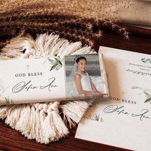 May include: A white chocolate bar with a floral design and the text "God Bless Sofia Ann". The bar is wrapped in clear plastic and features a photo of a young girl wearing a white dress.
