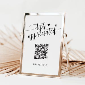 May include: A white sign with black text that reads "tips appreciated" with a small black heart symbol. A black and white QR code is below the text. The sign is in a silver frame and is sitting on a table.