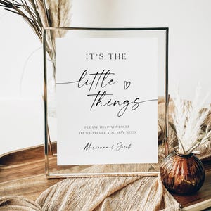 Puede incluir: Un cartel blanco con texto negro en un marco plateado. El cartel dice "It's the little things" con un pequeño símbolo de corazón. Debajo del texto se lee "Please help yourself to whatever you may need" y "Marunna & Jacob".
