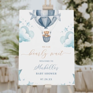May include: A blue and white hot air balloon with a teddy bear in the basket. The balloon is surrounded by clouds and stars. The text "We can barely wait" is written in cursive script. The text "Welcome to Michelle's Baby Shower" is written in blue. The date "07.28.XX" is written below the text.