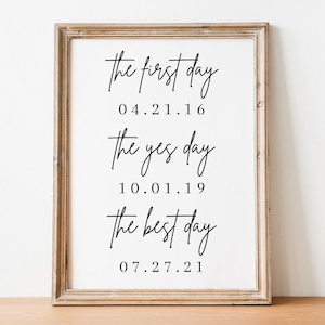 May include: A framed print with the text "the first day 04.21.16 the yes day 10.01.19 the best day 07.27.21" in black script font on a white background.