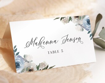 May include: White wedding place card with blue watercolour flowers and greenery. The text on the card reads "Makenna Jenson" and "Table 5".