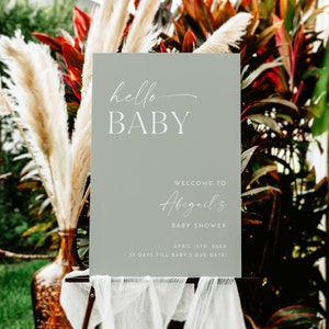 May include: A sage green welcome sign with white text that reads "hello BABY", "WELCOME TO Abigail's BABY SHOWER", "APRIL 1STH, 20XX", and "35 DAYS TILL BABY'S DUE DATE!"