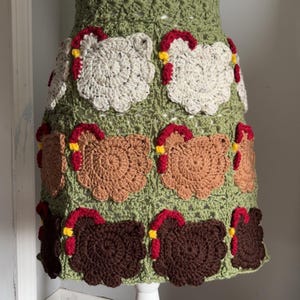 May include: A handmade crochet skirt featuring a repeating pattern of chickens. The skirt is primarily olive green with chicken motifs in white, brown, and dark brown, each with red and yellow accents. The skirt is displayed on a white mannequin.