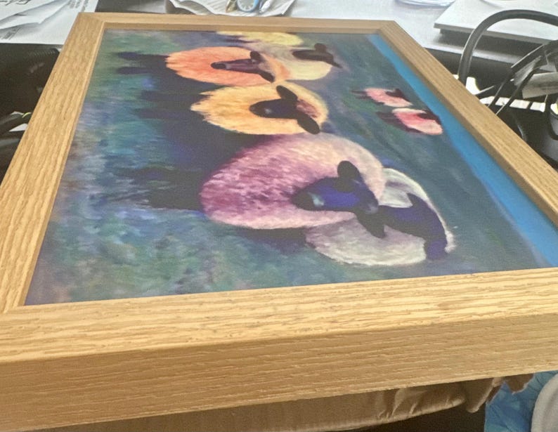 May include: Framed artwork featuring a watercolor painting of several swans floating on water. The swans have black heads and necks, with white and colorful bodies in shades of pink, orange, and yellow. The water is blue and green.