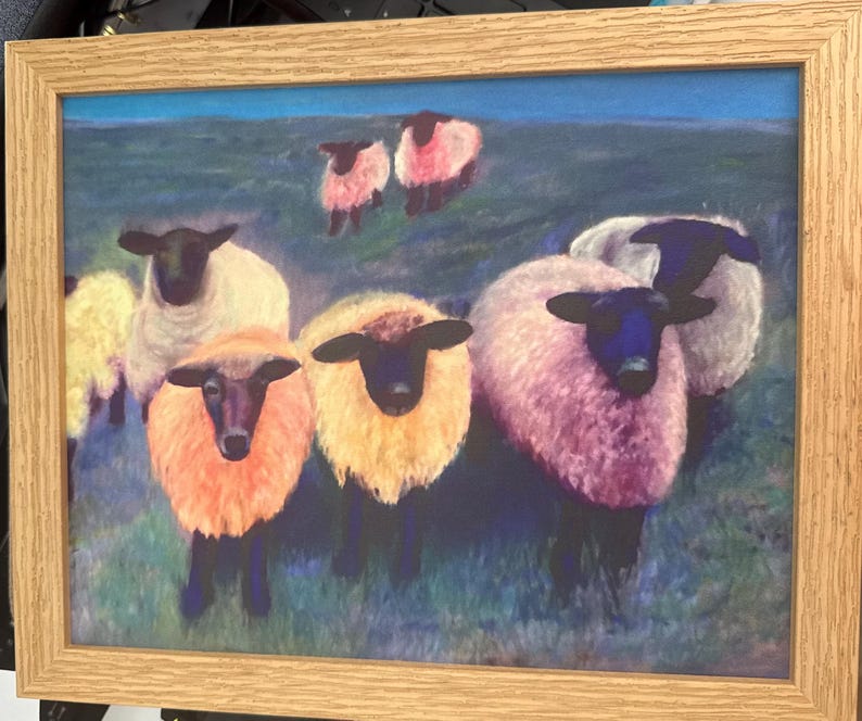 May include: A framed print featuring a group of colorful sheep. The sheep have black faces and legs, with wool in shades of pink, orange, yellow, and white. The background shows a green field and blue sky.