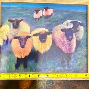 May include: A framed print featuring a flock of sheep with black faces and colorful wool. The sheep have wool in shades of yellow, orange, and pink. The background is a green field with a blue sky.