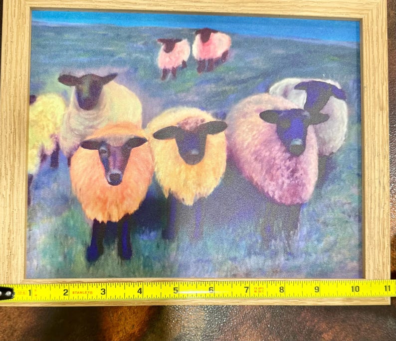 May include: A framed print featuring a flock of sheep with black faces and colorful wool. The sheep have wool in shades of yellow, orange, and pink. The background is a green field with a blue sky.