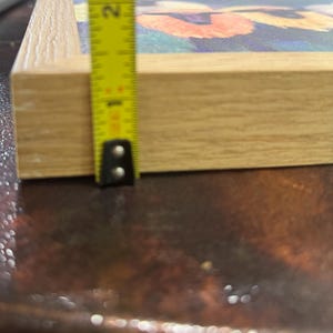 May include: A framed artwork with a floral design, featuring orange and yellow flowers against a blue background. The frame is made of light-colored wood. The artwork is approximately 1 inch thick, as measured by a yellow tape measure.