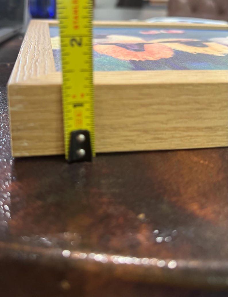 May include: A framed artwork with a floral design, featuring orange and yellow flowers against a blue background. The frame is made of light-colored wood. The artwork is approximately 1 inch thick, as measured by a yellow tape measure.