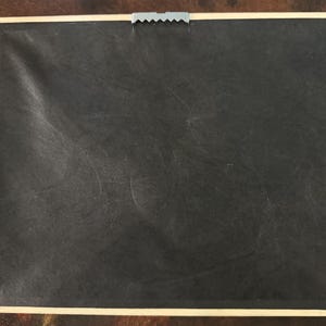 May include: Back of a framed artwork. The frame is a light beige color, and the backing is black. A metal sawtooth hanger is attached to the top center of the frame.