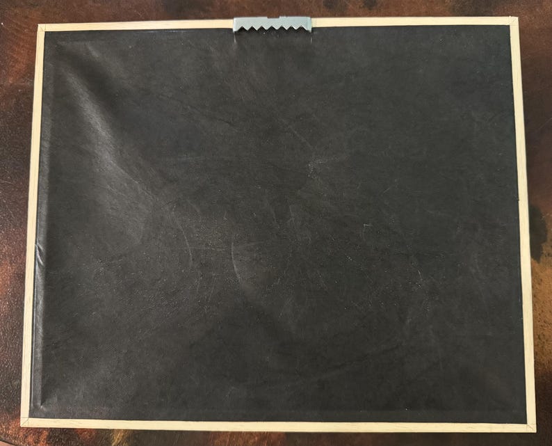 May include: Back of a framed artwork. The frame is a light beige color, and the backing is black. A metal sawtooth hanger is attached to the top center of the frame.