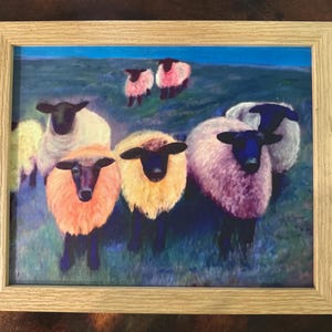 May include: Framed artwork featuring a flock of sheep in a field. The sheep have black faces and legs, with wool in shades of white, pink, and yellow. The background shows a blue sky and green grass. The frame is made of light-colored wood.