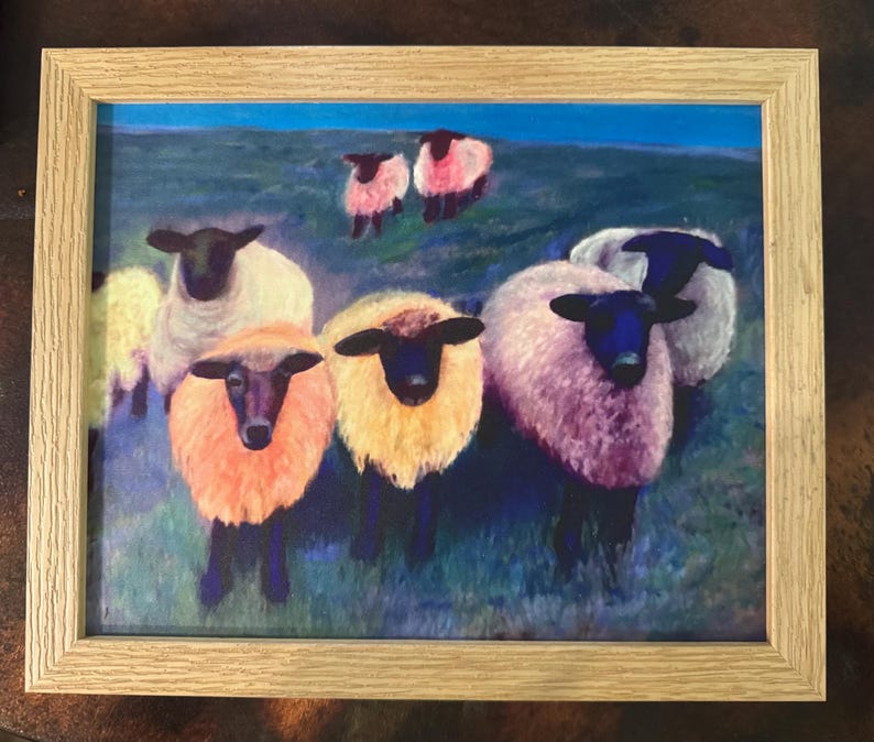 May include: Framed artwork featuring a flock of sheep in a field. The sheep have black faces and legs, with wool in shades of white, pink, and yellow. The background shows a blue sky and green grass. The frame is made of light-colored wood.