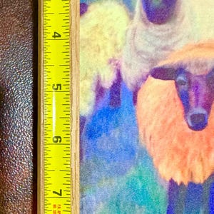 May include: A framed artwork featuring a colorful painting of sheep. The painting showcases sheep with white, orange, and yellow wool against a blue and green background. The frame is made of light-colored wood, and the artwork measures approximately 9 inches in height.