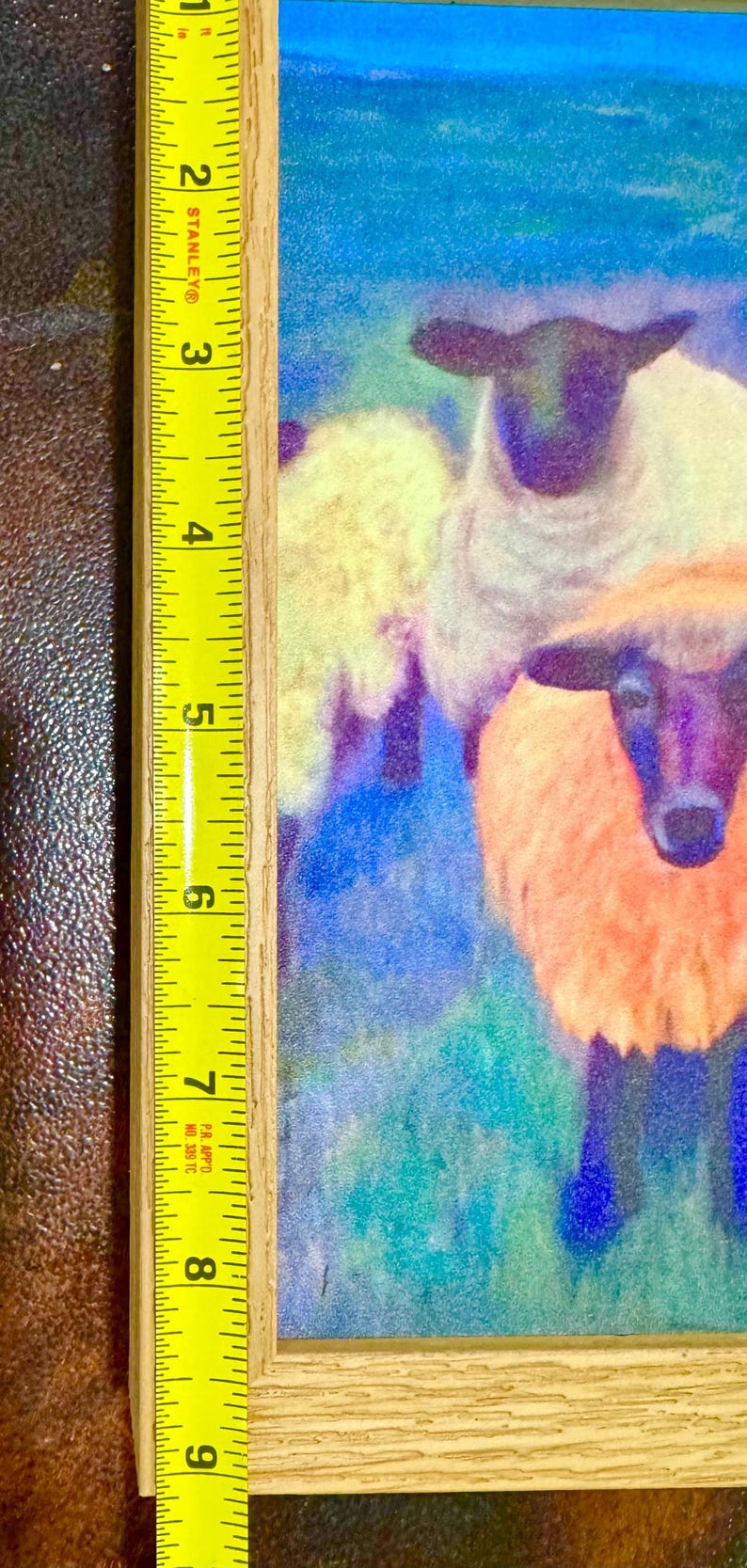 May include: A framed artwork featuring a colorful painting of sheep. The painting showcases sheep with white, orange, and yellow wool against a blue and green background. The frame is made of light-colored wood, and the artwork measures approximately 9 inches in height.