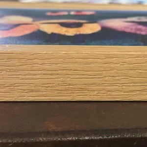 May include: A square, light-colored wooden picture frame. The frame surrounds a colorful image of flowers. The frame is made of light-colored wood with visible grain. The image is visible through the frame.