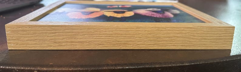 May include: A square, light-colored wooden picture frame. The frame surrounds a colorful image of flowers. The frame is made of light-colored wood with visible grain. The image is visible through the frame.