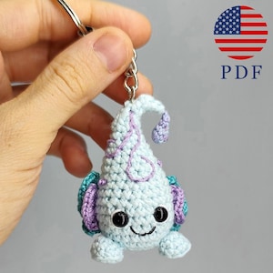 May include: A handmade crochet keychain in the shape of a whimsical creature. The keychain is primarily light blue with purple and teal accents. It has a smiling face with black eyes and a curved mouth. The keychain is attached to a silver metal keyring.