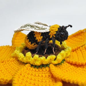 Puede incluir: Una abeja de ganchillo, a rayas negras y amarillas, posada sobre un girasol amarillo de ganchillo. La abeja tiene alas y detalles delicados. El girasol tiene m&uacute;ltiples capas de p&eacute;talos amarillos y un centro marr&oacute;n, todo hecho a mano.