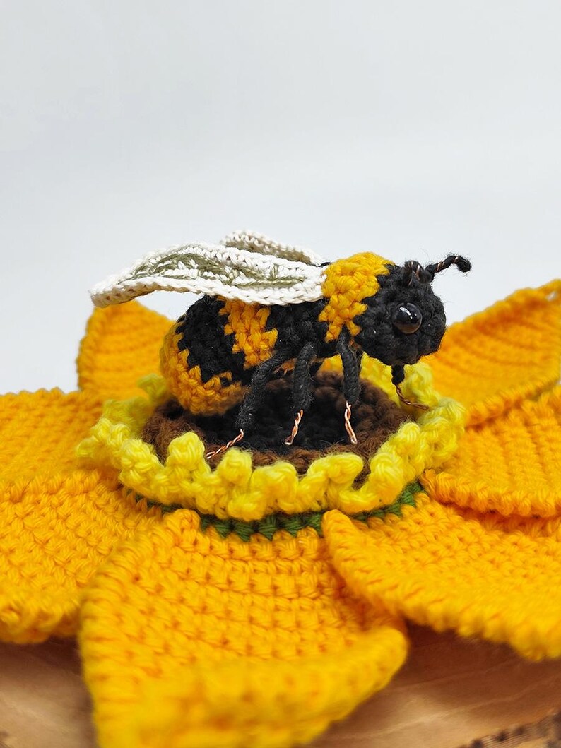 Puede incluir: Una abeja de ganchillo, a rayas negras y amarillas, posada sobre un girasol amarillo de ganchillo. La abeja tiene alas y detalles delicados. El girasol tiene m&uacute;ltiples capas de p&eacute;talos amarillos y un centro marr&oacute;n, todo hecho a mano.