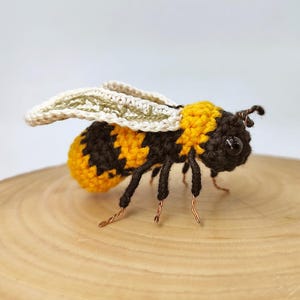 Puede incluir: Una abeja de ganchillo hecha a mano con segmentos corporales amarillos y negros, patas negras y delicadas alas. La abeja est&aacute; posada sobre una superficie de madera, mostrando los intrincados detalles del trabajo con hilo. Las antenas son de alambre.