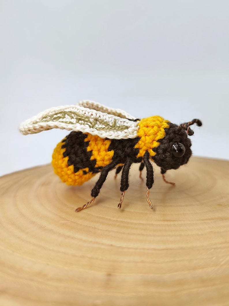 Puede incluir: Una abeja de ganchillo hecha a mano con segmentos corporales amarillos y negros, patas negras y delicadas alas. La abeja est&aacute; posada sobre una superficie de madera, mostrando los intrincados detalles del trabajo con hilo. Las antenas son de alambre.