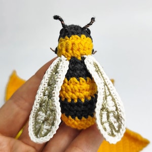 Puede incluir: Una abeja hecha a mano con ganchillo, con rayas negras y amarillas, alas blancas y antenas negras. Las alas tienen un patr&oacute;n detallado y est&aacute;n bordeadas en blanco. Un peque&ntilde;o objeto artesanal detallado, probablemente para decoraci&oacute;n o como juguete.