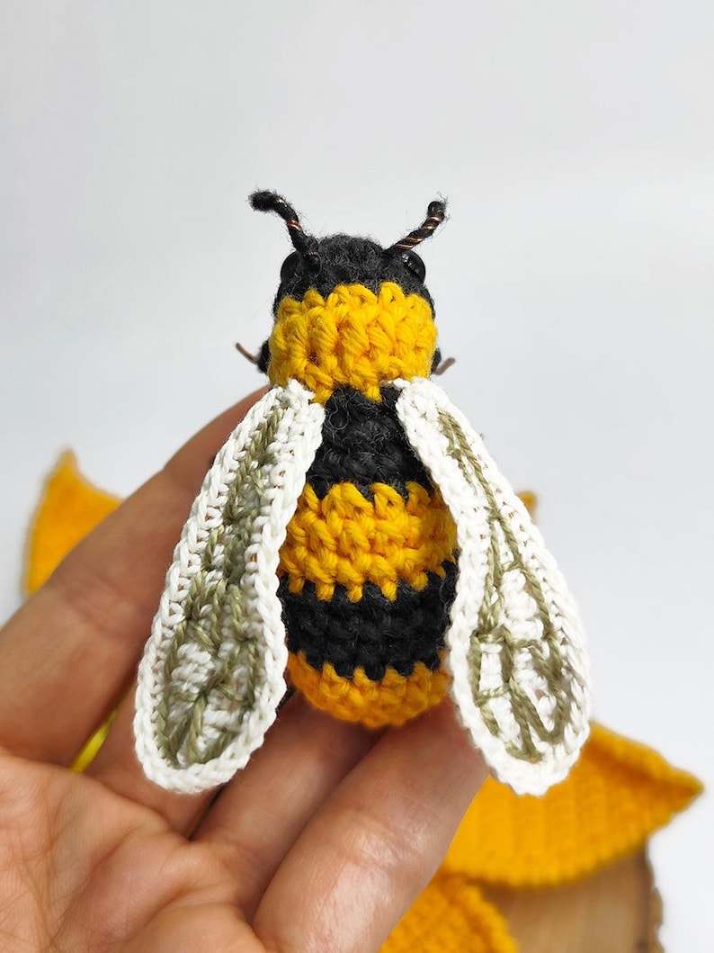 Puede incluir: Una abeja hecha a mano con ganchillo, con rayas negras y amarillas, alas blancas y antenas negras. Las alas tienen un patr&oacute;n detallado y est&aacute;n bordeadas en blanco. Un peque&ntilde;o objeto artesanal detallado, probablemente para decoraci&oacute;n o como juguete.