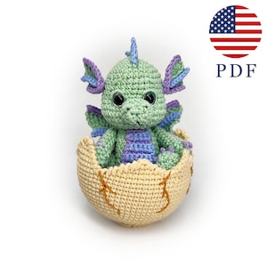 Könnte beinhalten: Ein gehäkelter Drache in einem cremefarbenen Ei. Der Drache ist grün mit lila und blauen Akzenten. Das Ei hat braune Nahtdetails. Das Bild enthält ein PDF-Symbol und eine amerikanische Flagge.