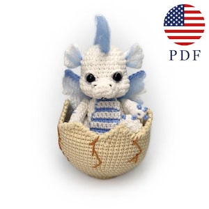 May include: A crocheted baby dragon in a cream-coloured egg. The dragon is white with blue accents on its wings, crest, and belly. The egg is beige with brown stitching. The image includes the text "PDF" and an American flag.