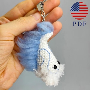 May include: A handmade keychain featuring a white and blue crocheted fish with black eyes and a fluffy tail. The keychain is attached to a silver metal ring. The image also includes a graphic of the American flag and the letters "PDF".