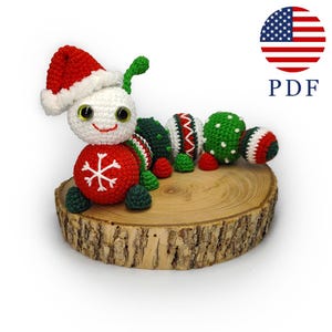Puede incluir: Un juguete de oruga navideña de ganchillo hecho a mano. La oruga tiene un cuerpo rojo con un diseño de copo de nieve blanco, segmentos verdes y blancos, y un gorro de Papá Noel. Está sobre una rodaja de madera.