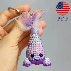 May include: A handmade crochet keychain in shades of purple, pink, and silver, featuring a whimsical creature design with black eyes and a smiling mouth. The keychain includes a silver metal ring and fluffy purple and white accents.