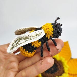Puede incluir: Una abeja de ganchillo hecha a mano con rayas amarillas y negras, alas blancas y patas de alambre. La abeja se sostiene sobre un girasol amarillo, tambi&eacute;n de ganchillo. Las antenas de la abeja son de alambre marr&oacute;n.