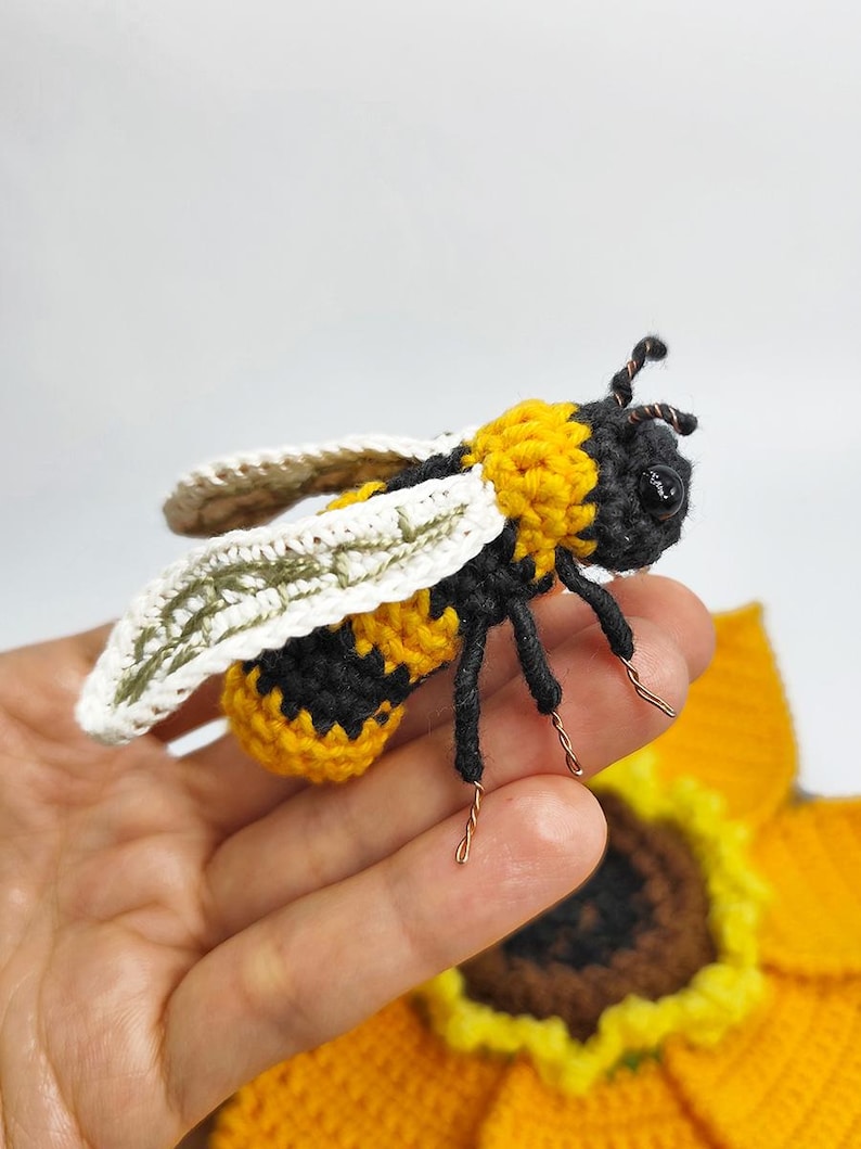 Puede incluir: Una abeja de ganchillo hecha a mano con rayas amarillas y negras, alas blancas y patas de alambre. La abeja se sostiene sobre un girasol amarillo, tambi&eacute;n de ganchillo. Las antenas de la abeja son de alambre marr&oacute;n.