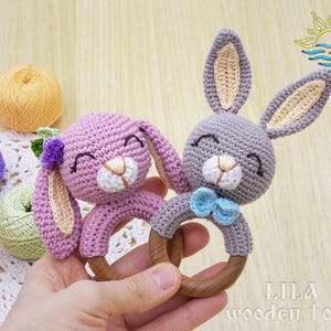 May include: A wooden teething ring with two crocheted bunny toys attached. One bunny is pink with a purple flower and the other is gray with a blue bow.