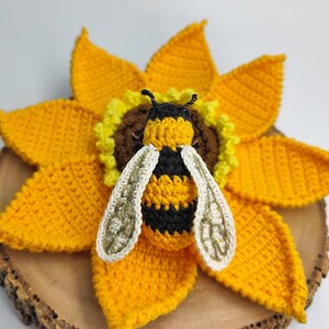 Puede incluir: Una abeja de ganchillo hecha a mano, con rayas negras y amarillas y alas blancas, posada sobre un girasol amarillo brillante. El girasol tiene un centro marr&oacute;n y est&aacute; colocado sobre una rodaja de madera.