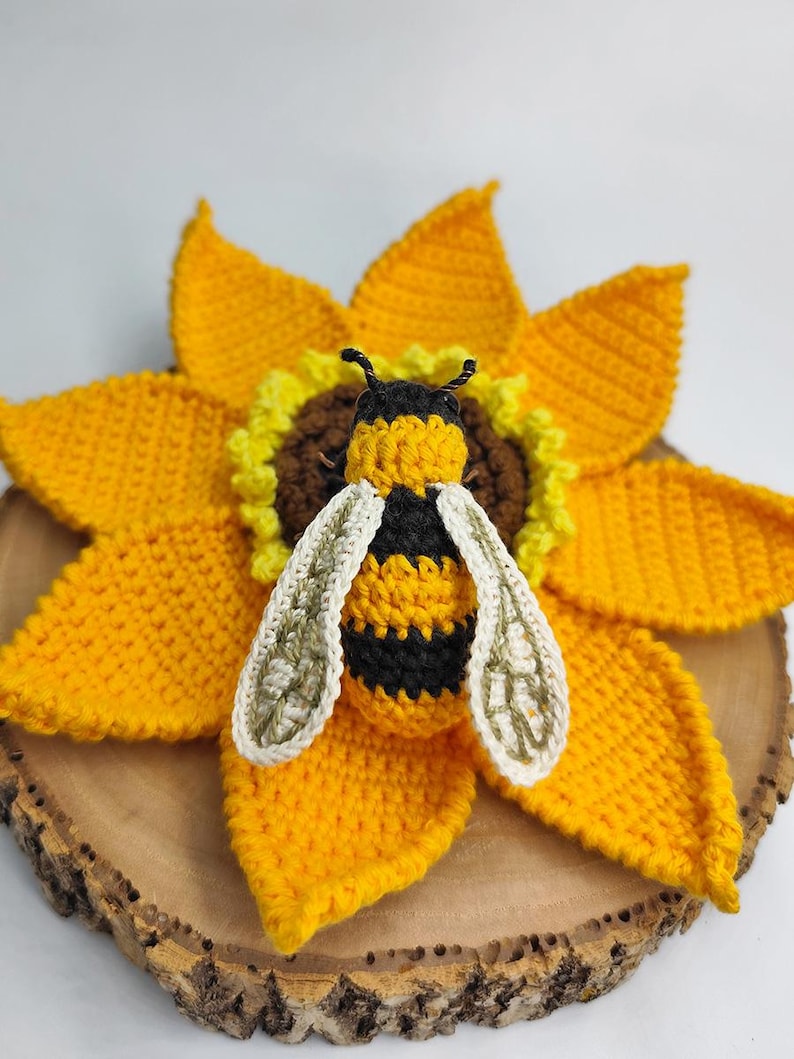Puede incluir: Una abeja de ganchillo hecha a mano, con rayas negras y amarillas y alas blancas, posada sobre un girasol amarillo brillante. El girasol tiene un centro marr&oacute;n y est&aacute; colocado sobre una rodaja de madera.