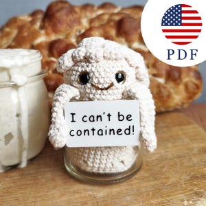 May include: A cream-colored crocheted creature with dark eyes and a small smile, sitting inside a clear glass jar. The creature holds a white rectangular sign with the black text "I can't be contained!" printed on the front surface.