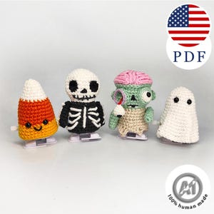 May include: Four crocheted wind-up toys featuring Halloween characters: a candy corn, a skeleton, a zombie with a pink brain and a dangling eyeball, and a white ghost. Each figure is mounted on a white plastic wind-up mechanism base.