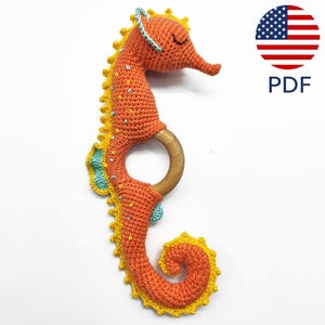 以下が含まれることがあります： 木製のかみかみリングに付けられた、オレンジ色の編みぐるみシーホース。黄色と青色のアクセントが施されています。PDF.