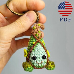May include: A handmade crochet keychain in the shape of a whimsical creature. The keychain features a light green body, a brown hat with green spots, and black button eyes. The design includes a smiling face and yellow accents. The keychain is attached to a bronze-colored ring.