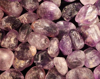 May include: A collection of tumbled amethyst stones featuring various shades of purple, ranging from light lavender to deep violet. Each stone has a smooth, polished surface with natural variations in clarity, internal fractures, and white inclusions.