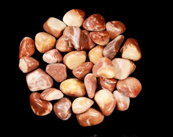 May include: A cluster of polished, tumbled stones in shades of brown, tan, and cream, set against a black backdrop. The stones vary in size and shape, with smooth, rounded surfaces. The overall aesthetic is natural and earthy.
