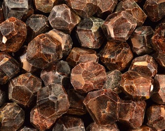 May include: A collection of dark red-brown garnet gemstones, each with multiple facets. The gemstones are piled together, showing their crystalline structure. The stones vary in size and shape, creating a textured appearance. The gemstones are a deep, rich colour.