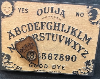 May include: A wooden Ouija board with a planchette in the shape of a heart. The board has the letters of the alphabet, numbers 1-9 and 0, the words "yes", "no", "goodbye", and "Ouija" printed on it. The planchette has the word "Ouija" and the number "3" printed on it.