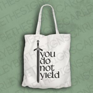 May include: White tote bag with a black sword graphic and the words "you do not yield". The bag has two handles and is made of a soft, opaque material. The design is centered on the front of the bag.
